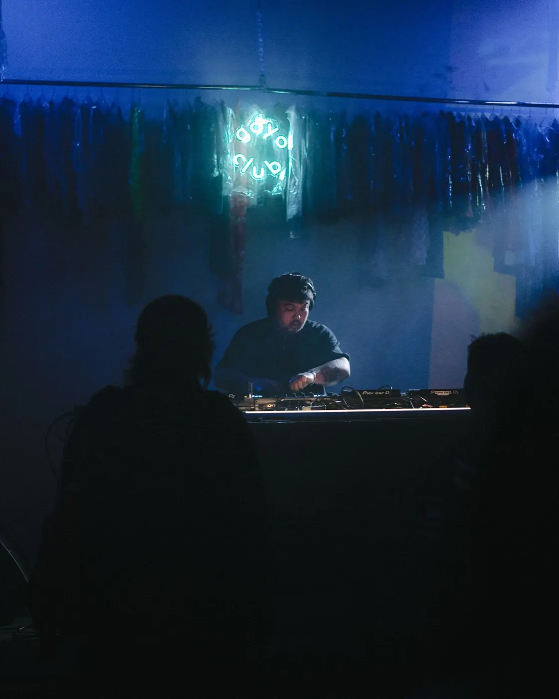DJ silhouetted behind the decks under the glowing goyo club sign, hanging garment racks in the background