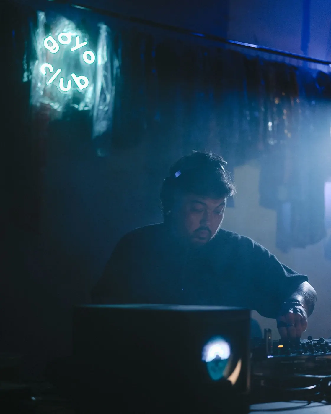 DJ in a cap focused on the mixer, alone at the booth in a haze of blue light