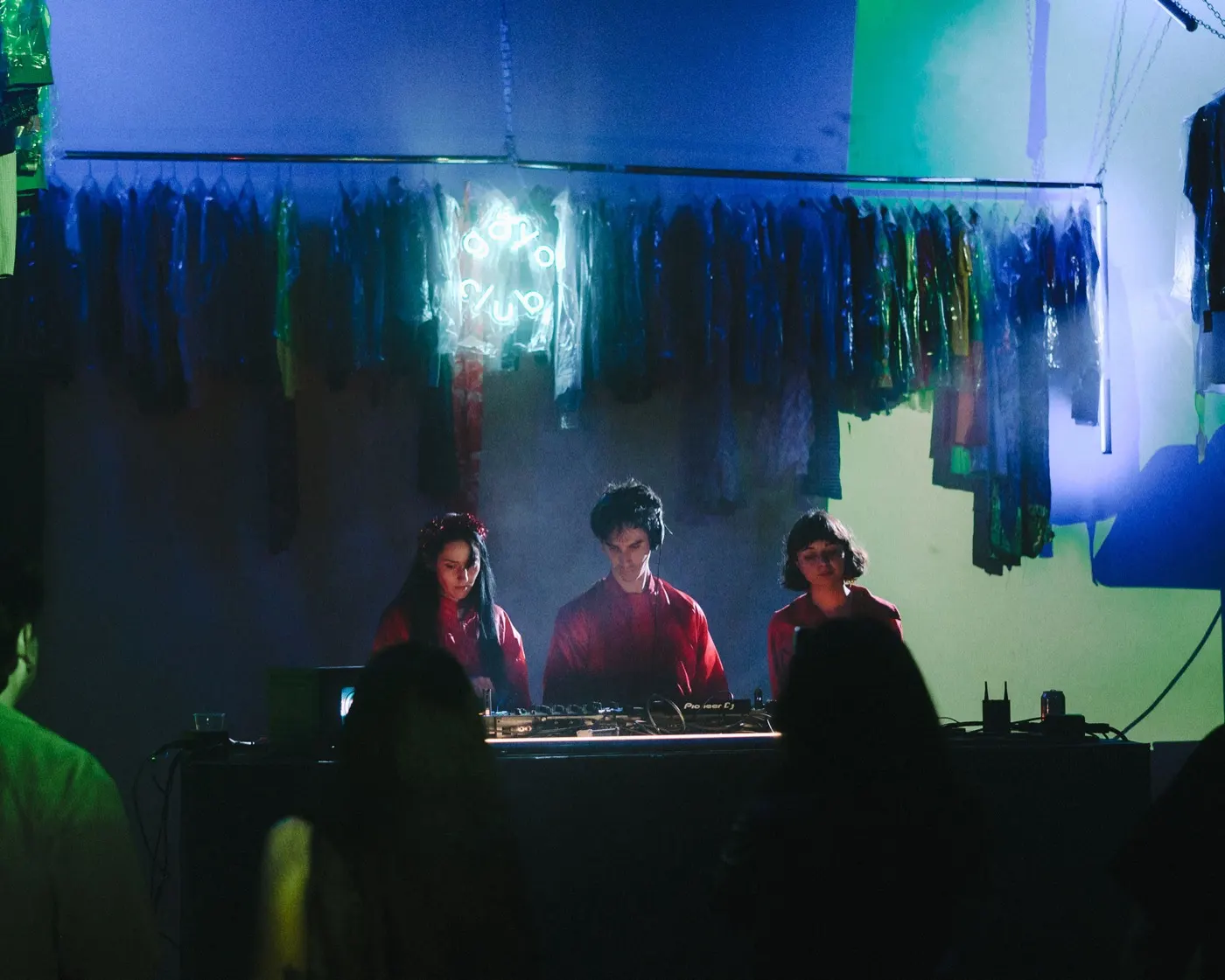 Three performers posed together behind the booth, lit in mixed red and green light