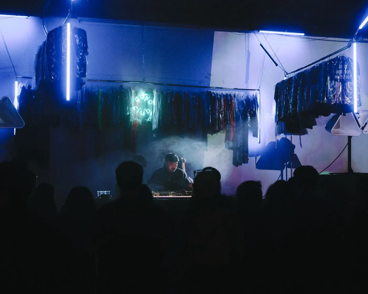 Wide view of the DJ booth beneath the goyo club neon sign, crowd silhouettes gathered in front of the garment racks