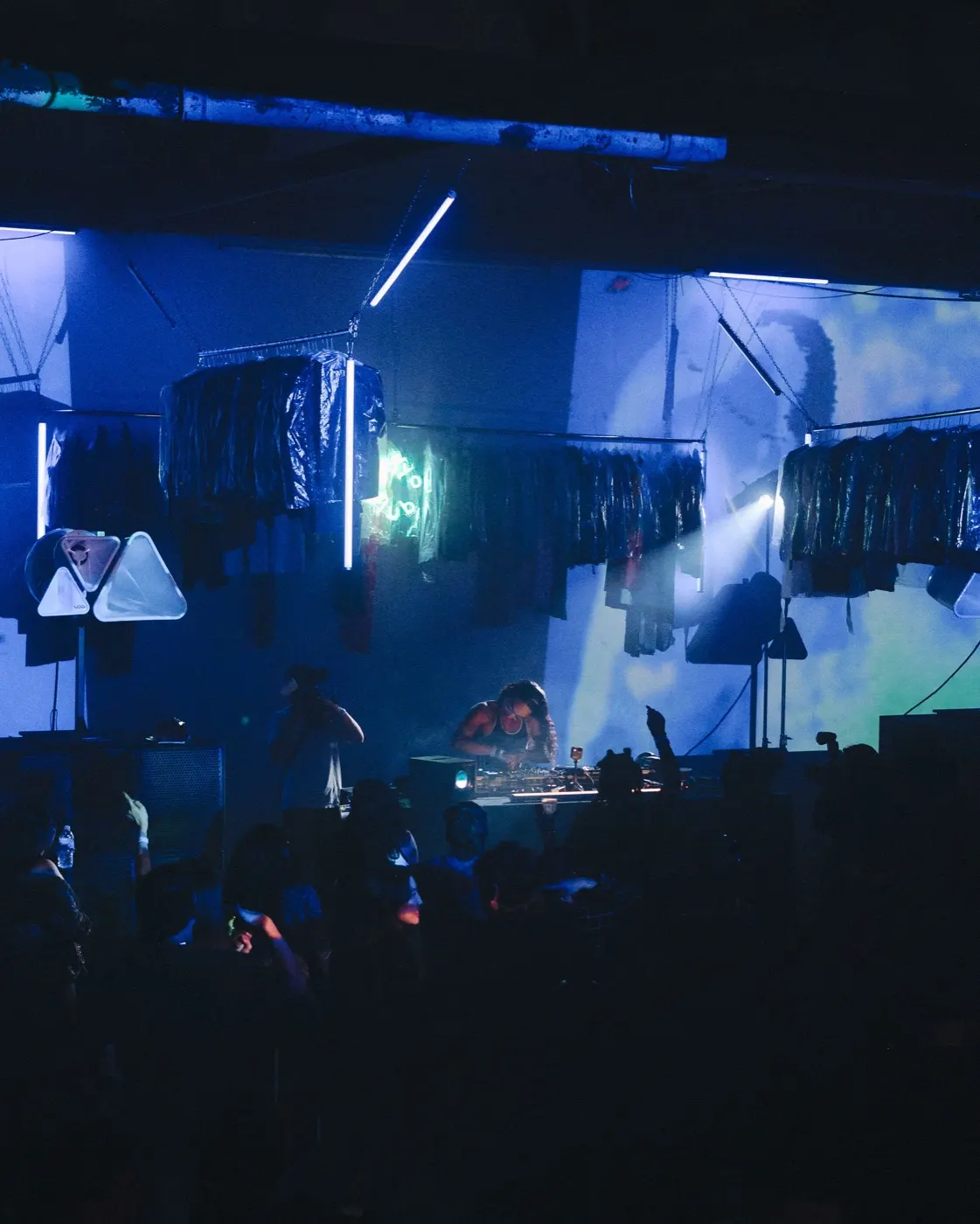 Atmospheric wide shot of the blue-lit dry cleaners racks with a small crowd in soft haze
