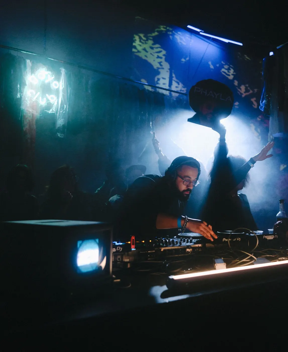 DJ silhouetted raising a hand energetically, warm string lights along the booth edge