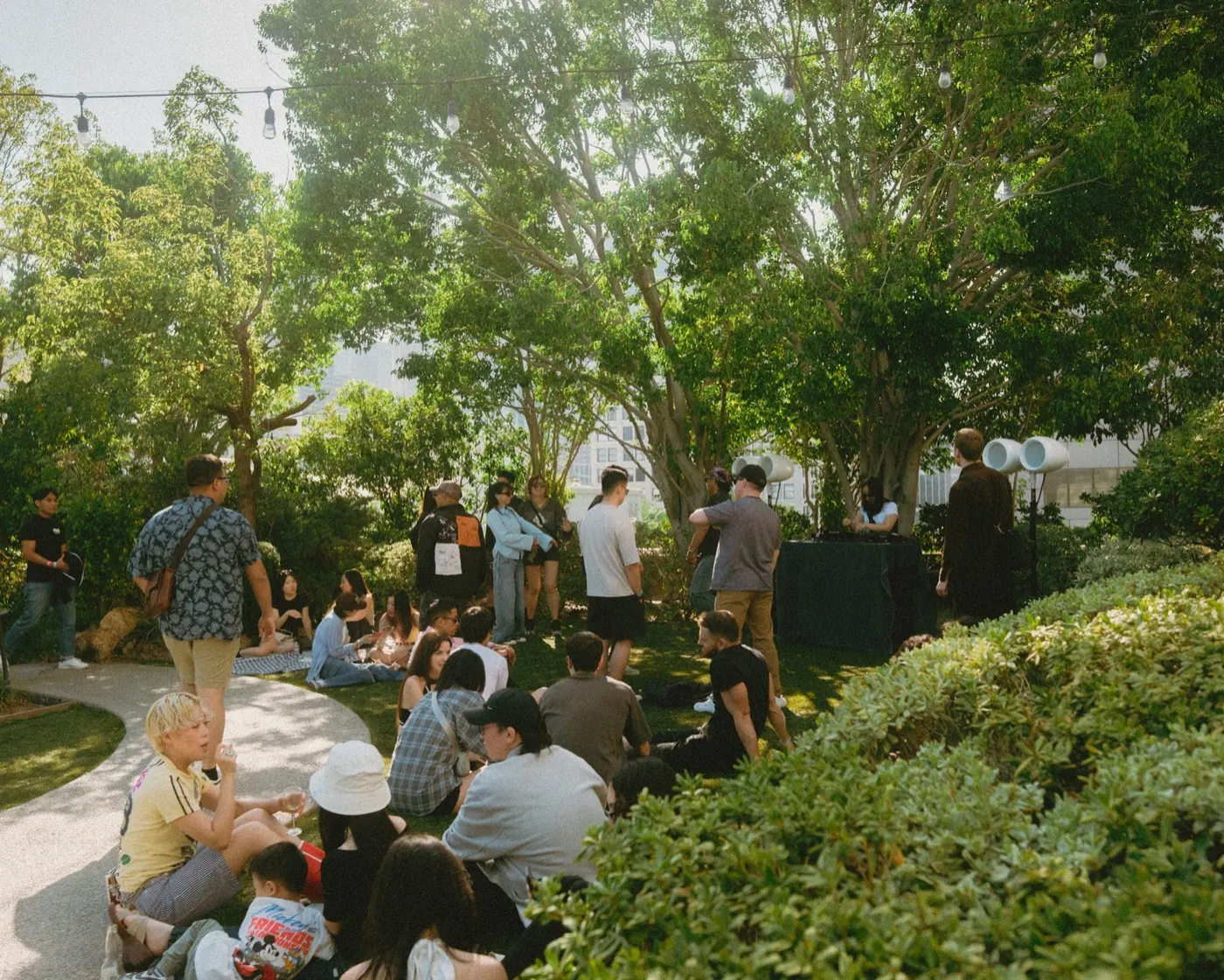Crowd seated and standing in the garden shade with the DJ setup visible behind them